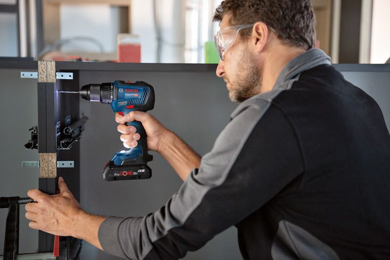 Een persoon die veiligheidsmateriaal draagt, bevestigt met een accuboormachine een plank aan een kast.