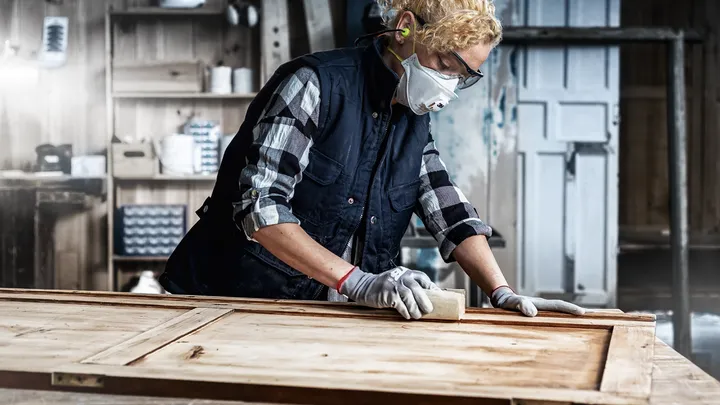 Een persoon met veiligheidsuitrusting schuurt een houten deur in een werkplaats.