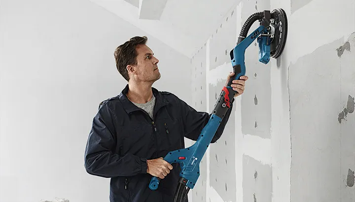 Iemand schuurt gipsplaat met een elektrische schuurmachine op een onafgewerkte muur.