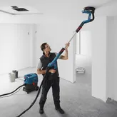 Een persoon met veiligheidsuitrusting schuurt een plafond met een gipsplaatschuurmachine die is aangesloten op een stofzuiger.