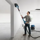 Iemand schuurt een witte muur met een schuurmachine voor gipsplaten die is aangesloten op een stofafzuiging.