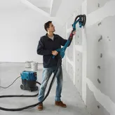 Iemand schuurt gipsplaat met een gipsplaatschuurmachine die is aangesloten op een stofafzuiging.