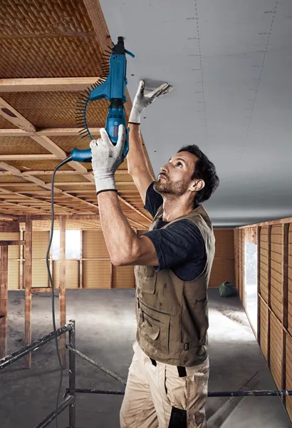 Een persoon met veiligheidsuitrusting monteert gipsplaat aan het plafond met behulp van een elektrische schroevendraaier.