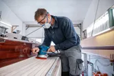 Een persoon met veiligheidsuitrusting schuurt een houten bootdek met behulp van een excentrische schuurmachine.