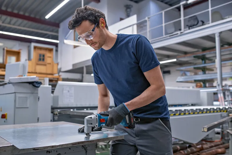 Een persoon die veiligheidsuitrusting draagt, zaagt een metalen plaat door met een elektrisch gereedschap in een werkplaats.