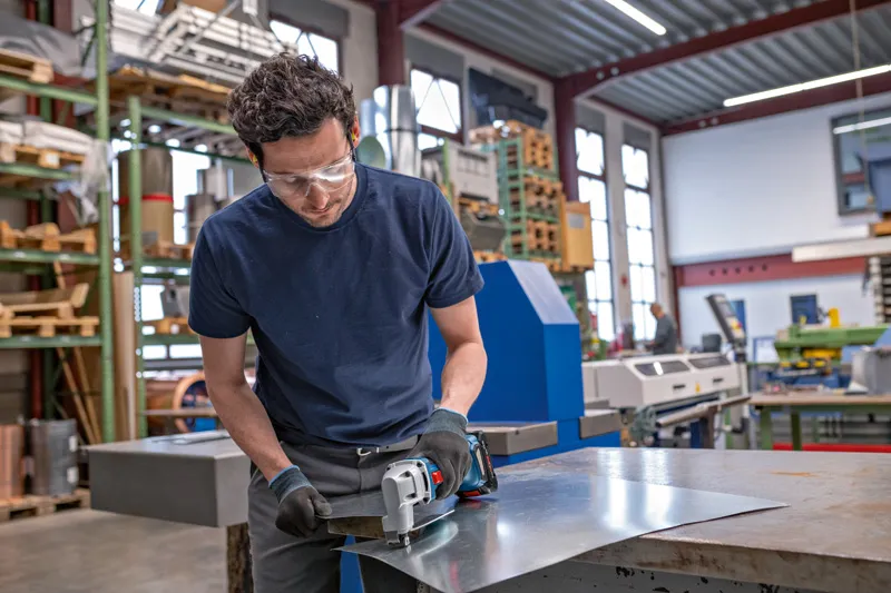 Een persoon die veiligheidsuitrusting draagt, snijdt plaatmetaal met een elektrisch gereedschap in een werkplaats.