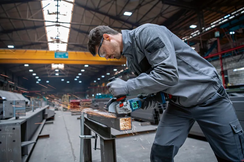 Een arbeider met veiligheidsuitrusting slijpt metaal in een industriële werkplaats.