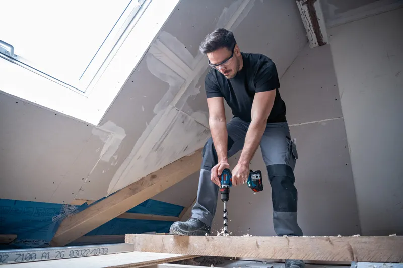 Een persoon met veiligheidsuitrusting boort in een houten balk op een gedeeltelijk afgewerkte zolder.
