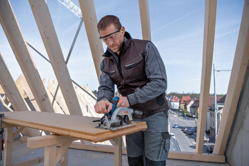 Een persoon met veiligheidsuitrusting zaagt op een bouwplaats een houten plank met een cirkelzaag.