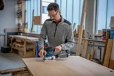 Een persoon met veiligheidsuitrusting schuurt een houten plank met een excentrische schuurmachine.