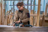 Een persoon met veiligheidsuitrusting gebruikt een excentrische schuurmachine op houten planken in een werkplaats.