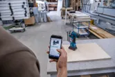 Iemand gebruikt een smartphone om een snoerloze boormachine in een houtbewerkingswerkplaats te raadplegen.