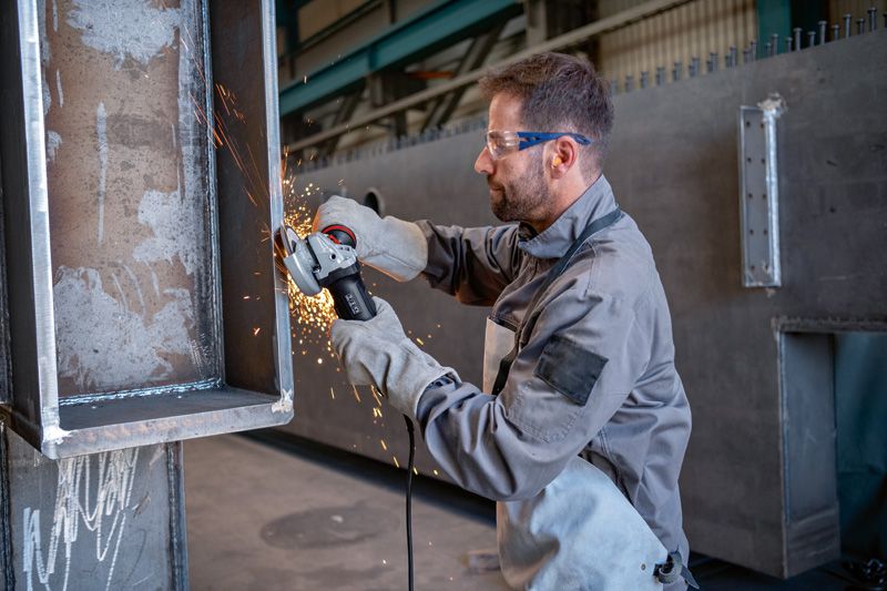 Een persoon met veiligheidsuitrusting slijpt de rand van een grote metalen balk.