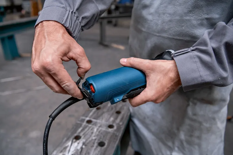 Persoon in werkkleding sluit een stroomkabel aan op een blauw handgereedschap.