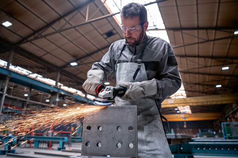 In een fabriek slijpt een persoon met veiligheidsuitrusting een metalen blok, waardoor vonken ontstaan.