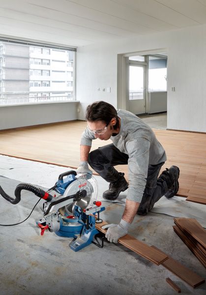 Een persoon met veiligheidsuitrusting zaagt een houten vloer met een verstekzaag.