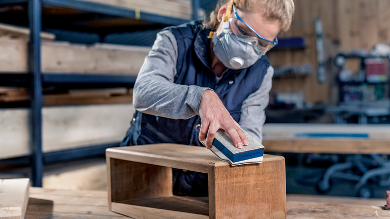 Een persoon met veiligheidsuitrusting schuurt een houten werkbank in een werkplaats.