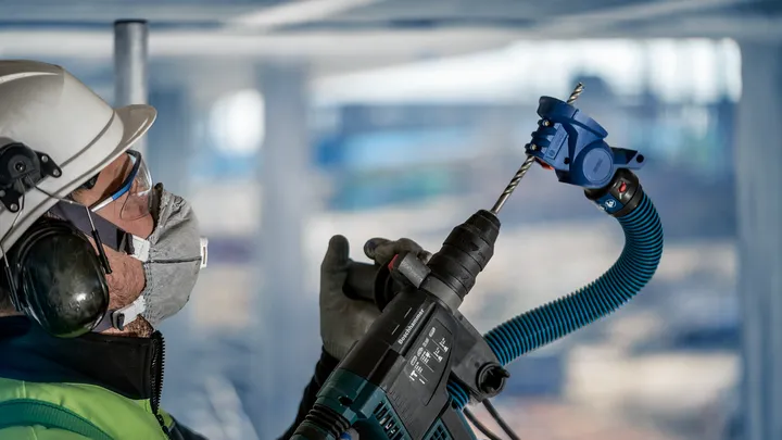 Een persoon met veiligheidsuitrusting boort met behulp van een elektrisch gereedschap met stofafzuiging in een plafond.