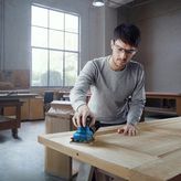 Een persoon met veiligheidsuitrusting schuurt een houten deur met een handschuurmachine.