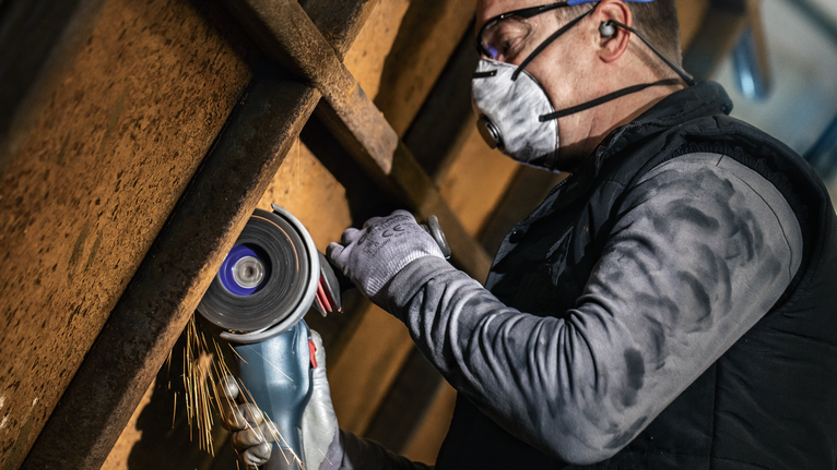Een persoon met veiligheidsuitrusting zaagt metaal met een handslijpmachine.