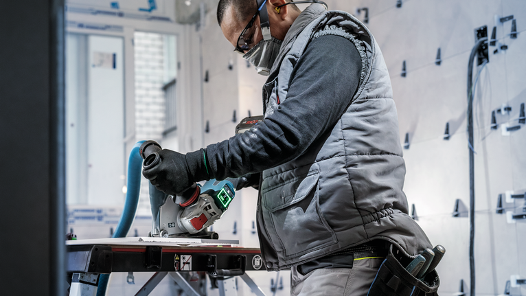 Een persoon die veiligheidsuitrusting draagt, gebruikt een elektrisch gereedschap om materiaal op een werkbank te snijden.