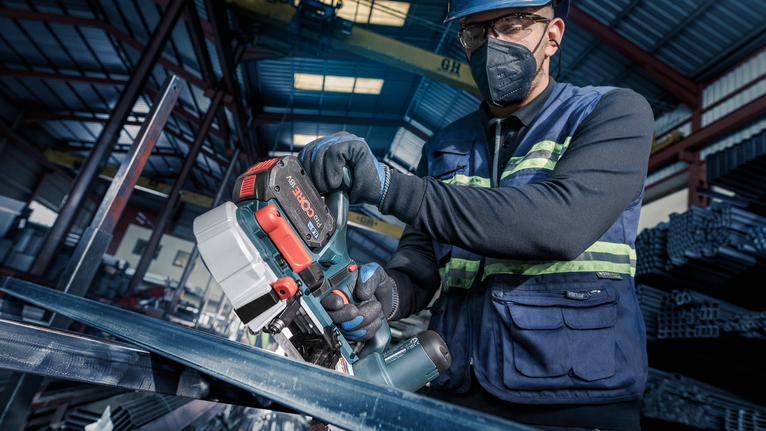 Een persoon met veiligheidsuitrusting zaagt metaal met een draagbare elektrische zaag in een industriële omgeving.