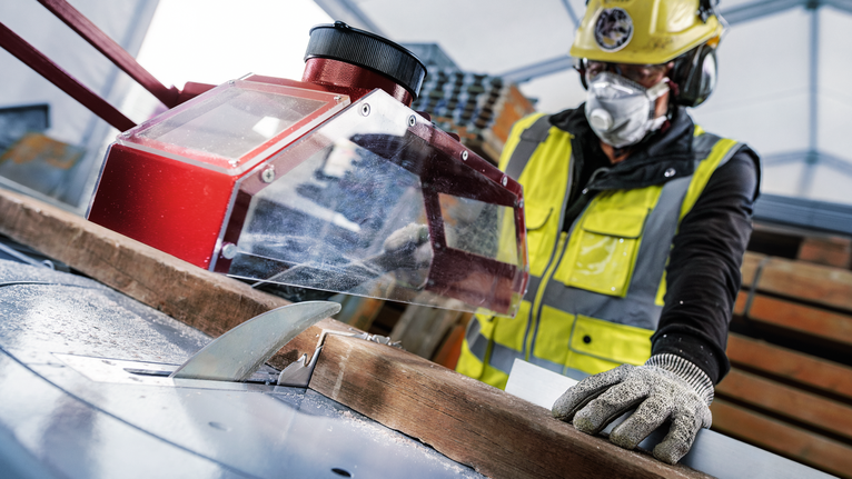 Een persoon met veiligheidsuitrusting geleidt hout door een tafelzaag.