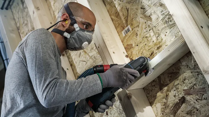 Een persoon met veiligheidsuitrusting schuurt een houten frame met een elektrisch gereedschap.