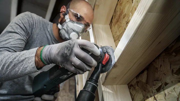 Een persoon die veiligheidsuitrusting draagt, gebruikt een haakse slijper om een houten frame te schuren.