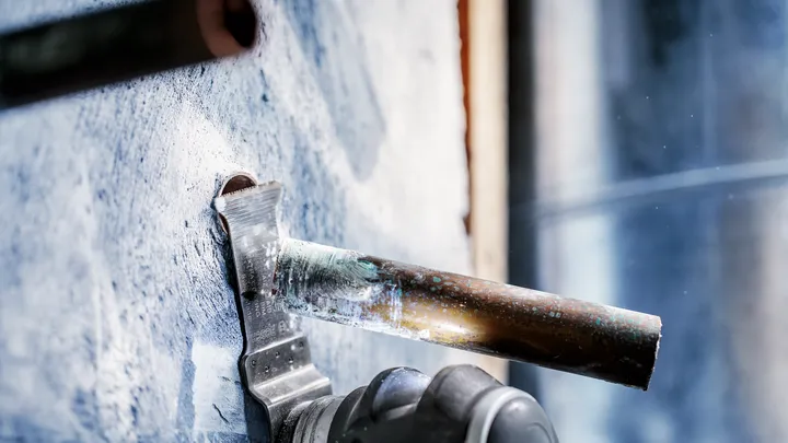 Elektrisch gereedschap zaagt door een metalen pijp die uit een betonnen muur steekt.