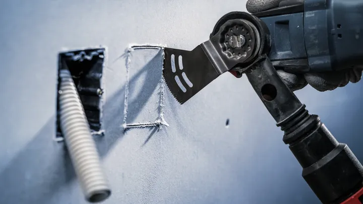 Een persoon met veiligheidsuitrusting maakt met een multitool een nette opening in de gipsplaat.