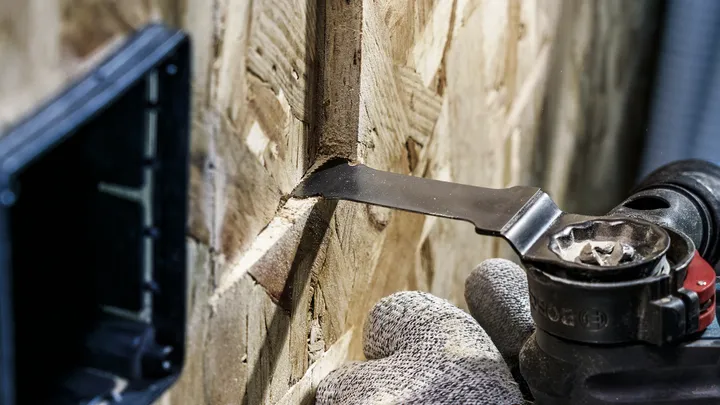 Een persoon die veiligheidsmateriaal draagt, gebruikt een multitool om een smalle sleuf in een houten paneel te zagen.