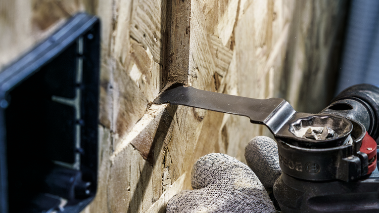 Een persoon die veiligheidsmateriaal draagt, gebruikt een multitool om een smalle sleuf in een houten paneel te zagen.