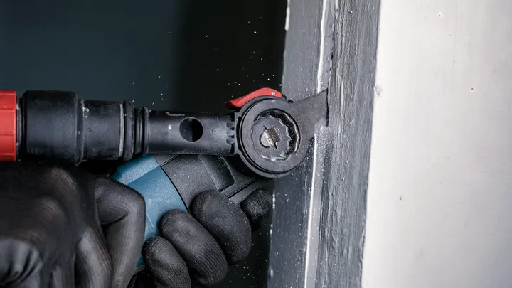 Een persoon met veiligheidsuitrusting schuurt een raamkozijn met een elektrisch gereedschap.