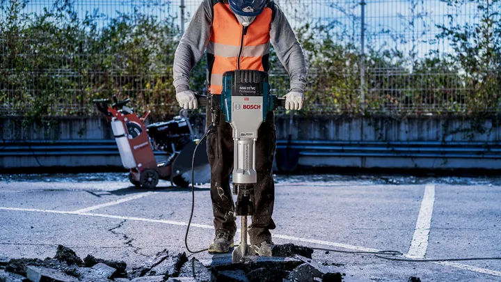 Een persoon met veiligheidsuitrusting gebruikt een breekhamer om het betonnen wegdek te breken.