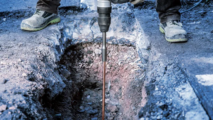 Een persoon met veiligheidsuitrusting boort met een zwaar elektrisch gereedschap in rotsachtige grond.