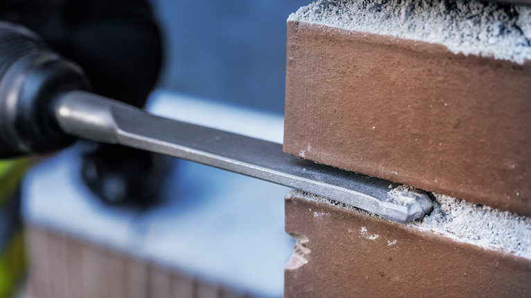 Een persoon die veiligheidsmateriaal draagt, gebruikt een beitel om een bakstenen muur te doorbreken.