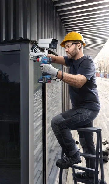 Een persoon die veiligheidsuitrusting draagt, installeert een beveiligingscamera met behulp van een boormachine.