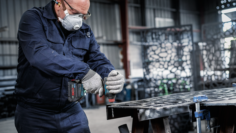 Een persoon met veiligheidsuitrusting boort in een metalen plaat in een werkplaats.