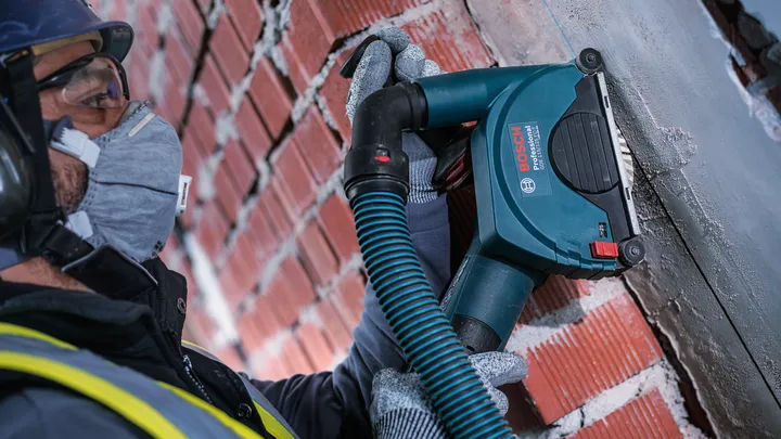 Een persoon met veiligheidsuitrusting slijpt met een elektrisch gereedschap de voegen van metselwerk in een bakstenen muur.