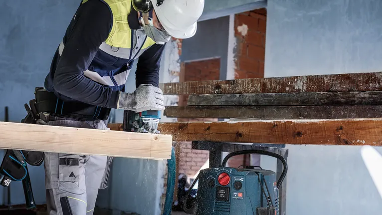 Een persoon met veiligheidsuitrusting boort in een houten balk op een bouwplaats.
