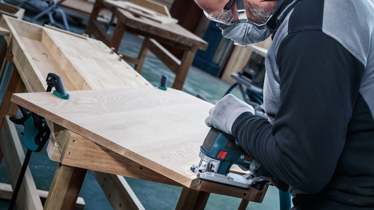 Een persoon die veiligheidsuitrusting draagt, gebruikt een decoupeerzaag om een houten plank te zagen.