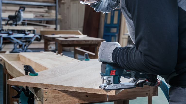 Een persoon met veiligheidsuitrusting zaagt hout met een decoupeerzaag in een werkplaats.