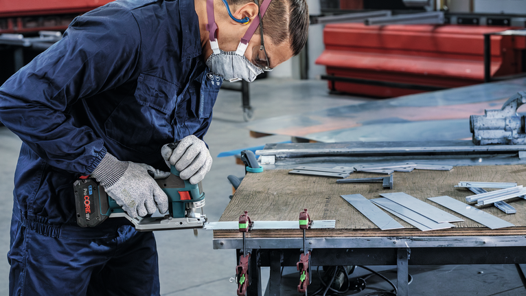 Een persoon met veiligheidsuitrusting zaagt metalen platen met een elektrische zaag op een werkbank.