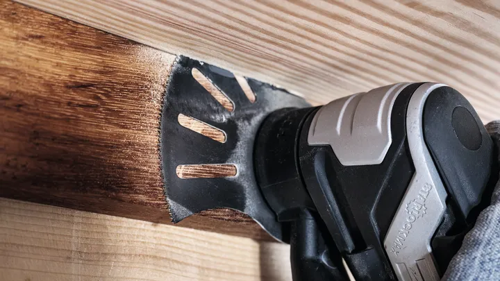 Een persoon met veiligheidsuitrusting gebruikt een multitool om houten balken te schuren.