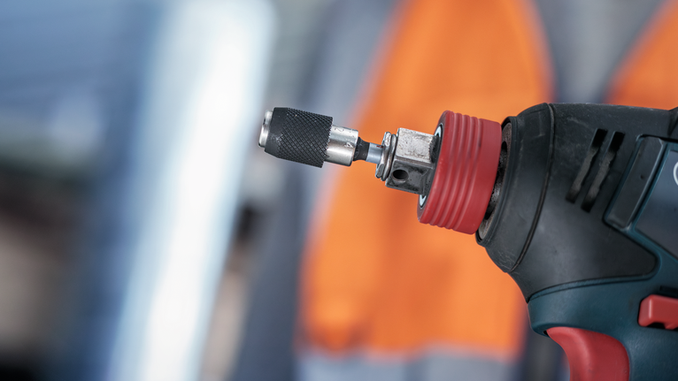 Er is een elektrisch gereedschap met een metaalboor te zien. Op de achtergrond draagt een persoon veiligheidsuitrusting.