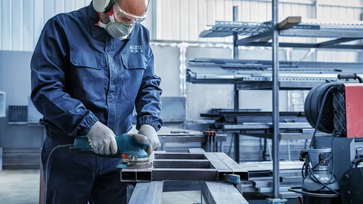Een persoon met veiligheidsuitrusting slijpt een metalen balk in een industriële werkplaats.