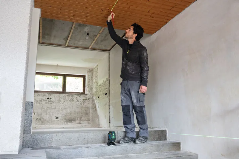 Iemand meet een plafond met een rolmaat, geleid door een laserwaterpasinstrument.