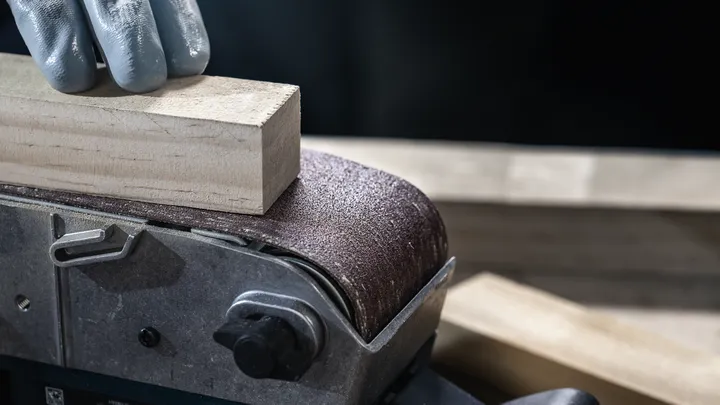 Een persoon die veiligheidsuitrusting draagt, schuurt een houten blok met een bandschuurmachine.