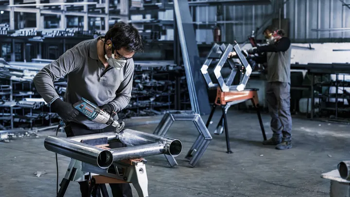 Een persoon met veiligheidsuitrusting boort in een metalen pijpconstructie in een werkplaats.